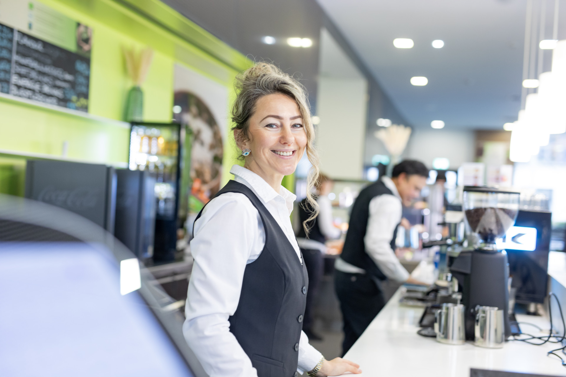 Gerda_Cafeteria_Cube Eine Frau in einem Café hinter einer Theke, lächelnd.